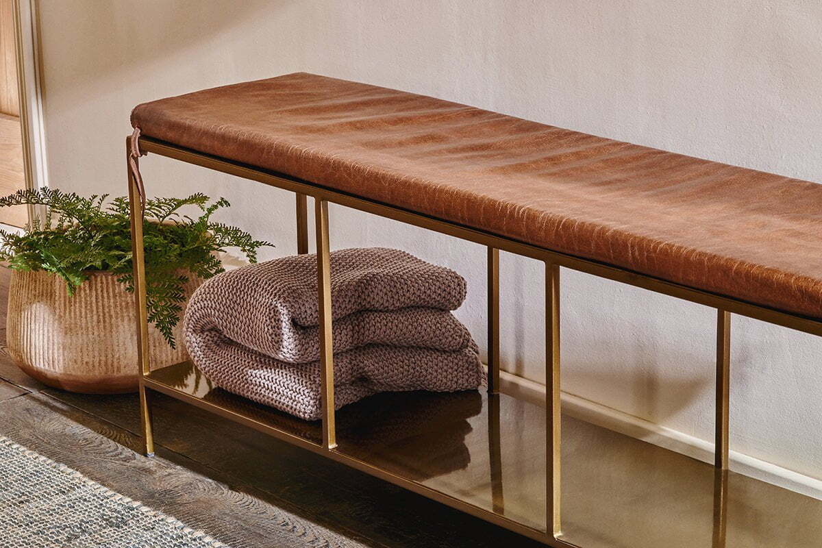The nkuku Mahi Iron & Leather Bench, featuring a brown leather cushion and brass frame, rests against the wall. On its lower shelf, two folded chunky knit gray blankets sit next to a woven basket with green ferns.
