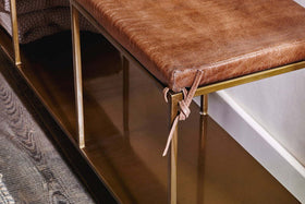 A close-up of the nkuku Mahi Iron & Leather Bench shows a brown leather cushion with thin ties, a brass frame, lower metal shelf, dark wood flooring, and a textured rug partially visible on the left.