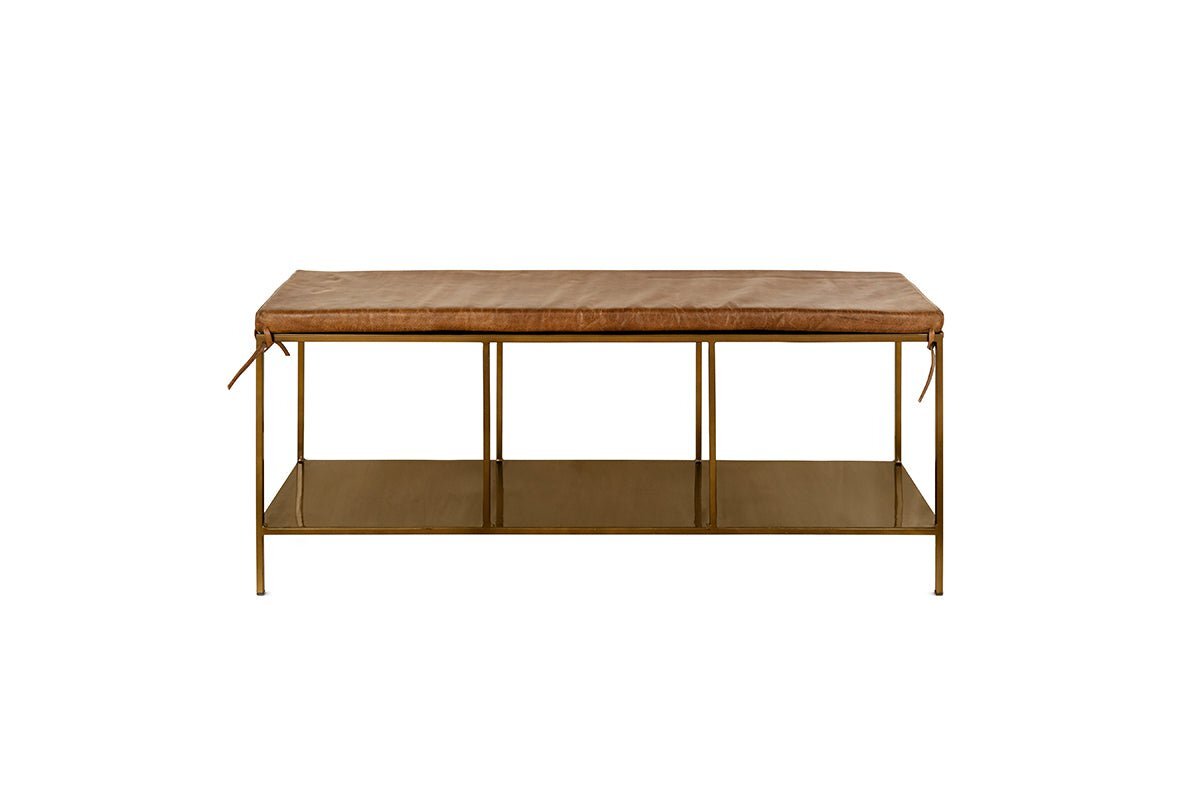 The nkuku Mahi Iron & Leather Bench features a rectangular tan leather cushion with side ties, slender brass legs, and a metal lower shelf. This minimalist bench is photographed against a plain white background.