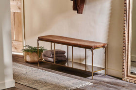 The nkuku Mahi Iron & Leather Bench with a tan leather seat and brass frame sits beneath a large mirror. A gray blanket is draped on top, while a basket with greenery is below. A textured rug softens the wood floor against the light wall.