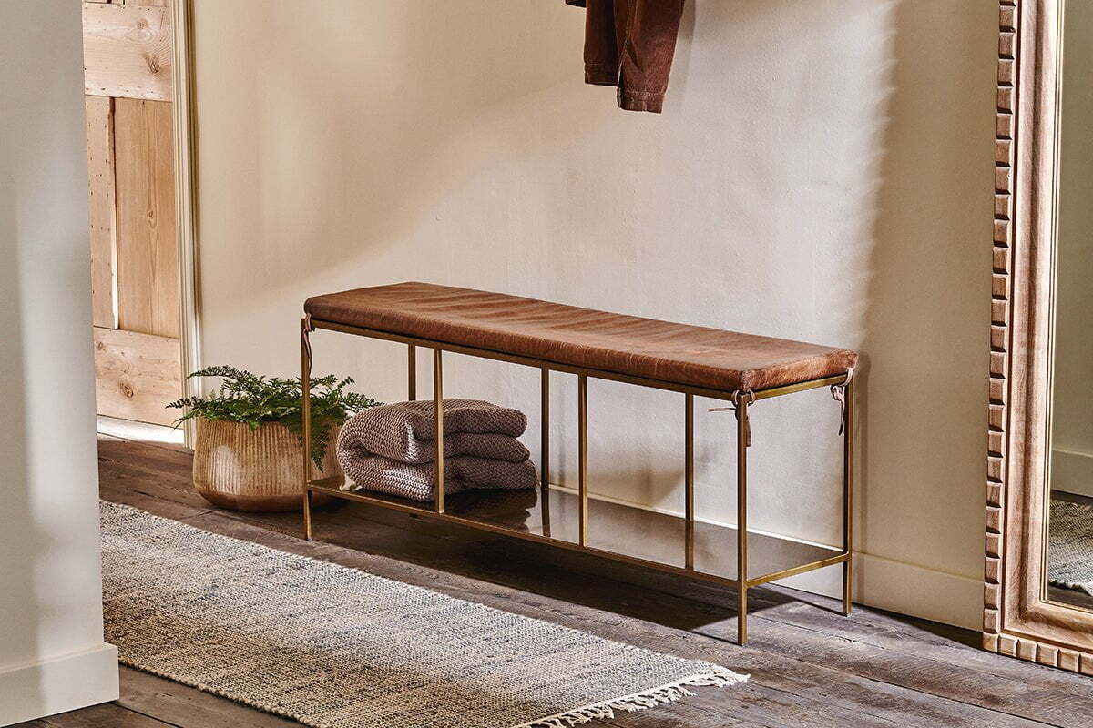 The nkuku Mahi Iron & Leather Bench with a tan leather seat and brass frame sits beneath a large mirror. A gray blanket is draped on top, while a basket with greenery is below. A textured rug softens the wood floor against the light wall.