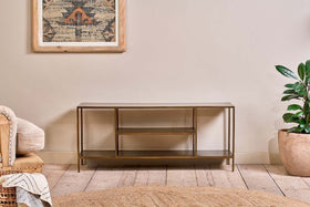 The nkuku Mahi Media Standing Shelf, with gold metal and brass shelves, stands against a beige wall. A potted plant is on the right, a cream armchair with a throw on the left, and a geometric tapestry hangs above on the wooden floor.