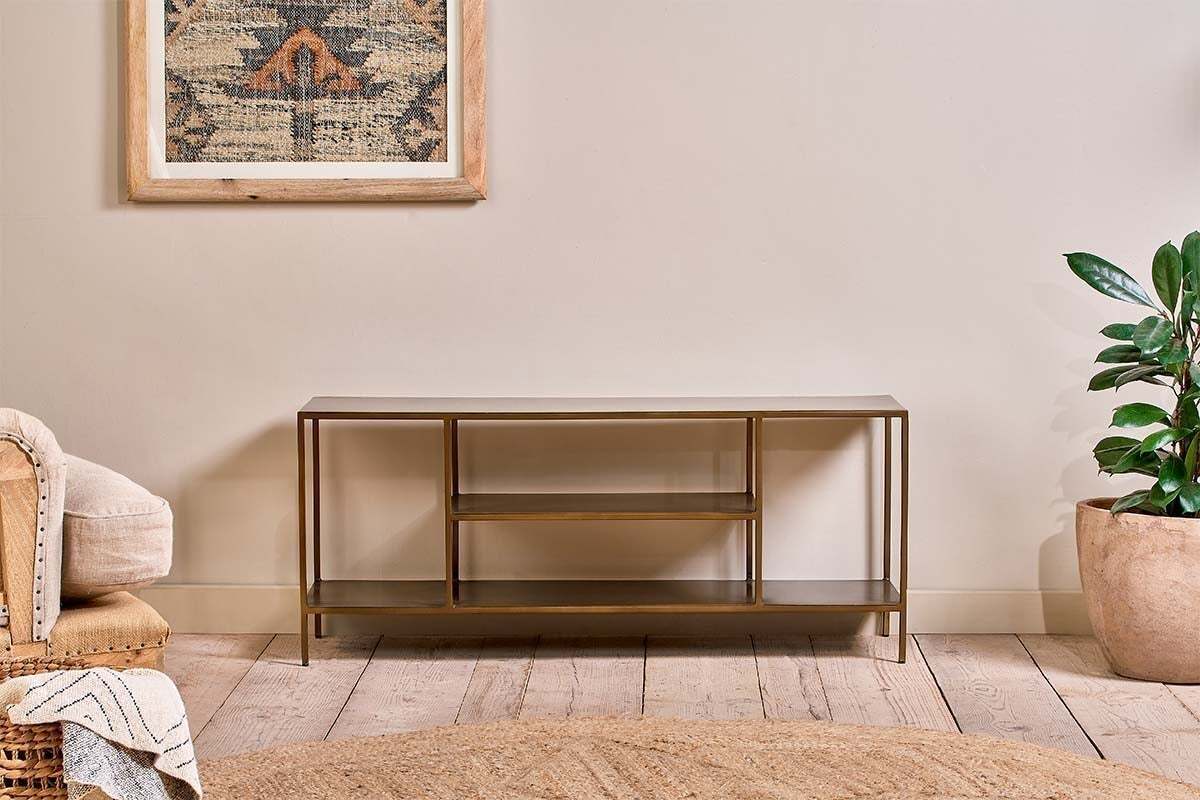 The nkuku Mahi Media Standing Shelf, with gold metal and brass shelves, stands against a beige wall. A potted plant is on the right, a cream armchair with a throw on the left, and a geometric tapestry hangs above on the wooden floor.