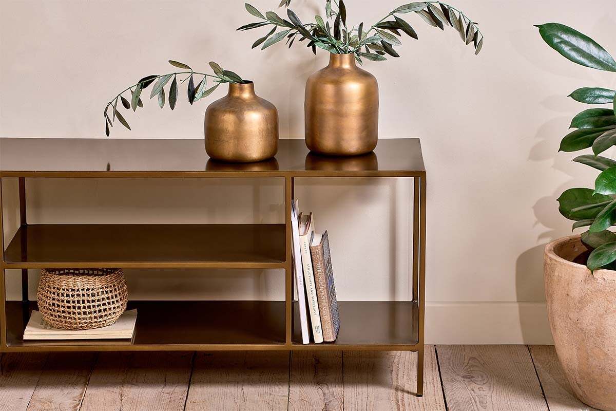The Mahi Media Standing Shelf by nkuku, featuring a bronze finish, displays two gold vases with leafy branches, books, and a small woven basket. To the right on the wooden floor sits a potted green plant against a cream-colored wall.