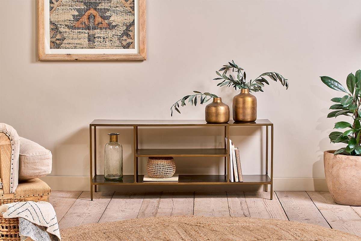 A modern living room features a gold metal console table with two gold vases, books, and a basket. Nearby, the nkuku Mahi Media Standing Shelf displays entertainment equipment, while plants and a cozy beige chair sit on light wood floors.
