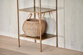 A woven basket with two handles sits on the bottom shelf of the nkuku Mahi Shelving Unit - Narrow, a minimalist brass piece placed against a light textured wall and wooden floor for stylish storage display.