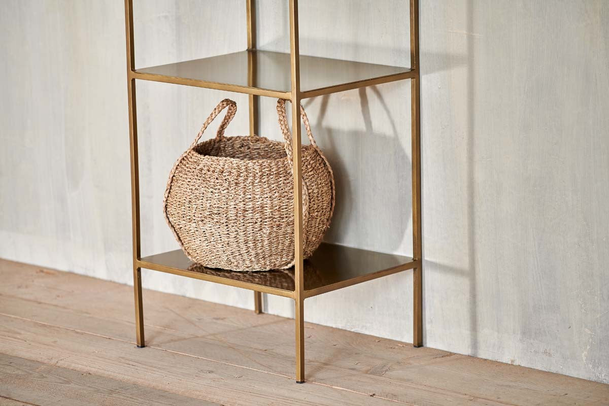 A woven basket with two handles sits on the bottom shelf of the nkuku Mahi Shelving Unit - Narrow, a minimalist brass piece placed against a light textured wall and wooden floor for stylish storage display.