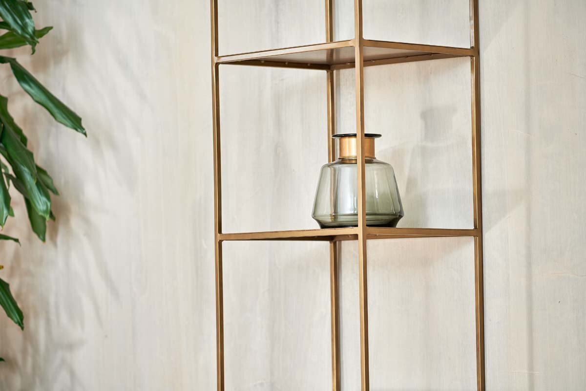 A glass jar with a dark lid rests on the nkuku Mahi Shelving Unit - Narrow, its sleek brass shelves contrasting against a light wall. A leafy green plant peeks in from the left, enhancing the minimalist and contemporary vibe.