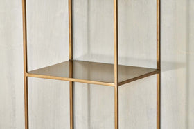 A close-up of the empty, gold metal Mahi Shelving Unit - Narrow by nkuku, featuring a square frame and open design, placed against a light gray textured wall. This brass shelf offers minimalist lines and modern, practical storage.