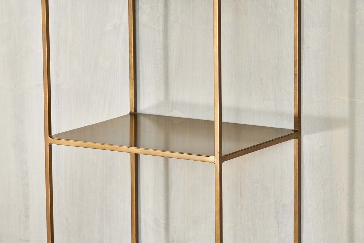 A close-up of the empty, gold metal Mahi Shelving Unit - Narrow by nkuku, featuring a square frame and open design, placed against a light gray textured wall. This brass shelf offers minimalist lines and modern, practical storage.