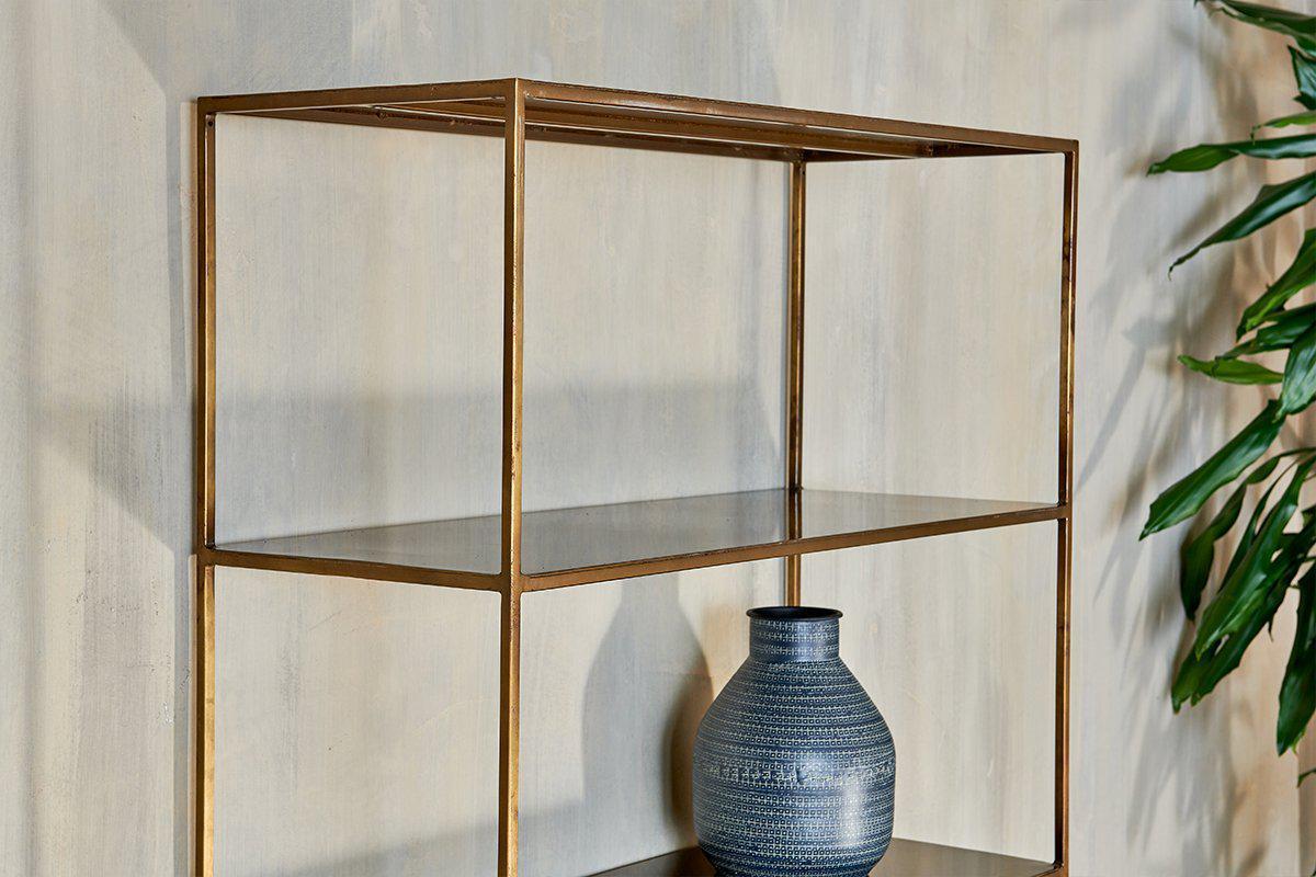 A minimalist nkuku Mahi Shelving Unit - Wide with a brass frame stands against a beige textured wall. On the middle shelf is a dark blue patterned ceramic vase, and a leafy green plant is partially visible on the right.