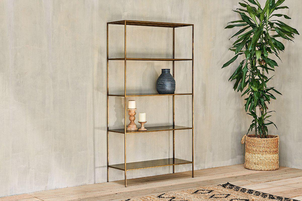 The nkuku Mahi Shelving Unit - Wide, featuring four brass and glass shelves, displays a blue vase, wood candlesticks, and a ceramic cup against a beige wall. A tall leafy plant in a basket stands nearby on the light wood floor.