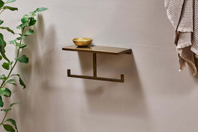 The nkuku Mahi Wall Mounted Toilet Roll Holder, featuring a bronze-colored brass shelf with two hooks and a small brass bowl on top, is fixed to a beige wall. A leafy plant rises on the left and a textured beige towel hangs on the right.
