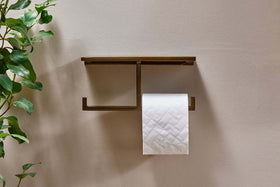 The nkuku Mahi Wall Mounted Toilet Roll Holder is fixed to a beige wall, holding a roll of toilet paper on its lower bar. A shelf sits above, with leafy green plant leaves visible on the left side of the image.