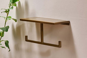 The Mahi Wall Mounted Toilet Roll Holder by nkuku, a small rectangular brass piece with a flat top and two-hook bar, is fixed to a beige wall; a green leafy plant appears partially on the left side.