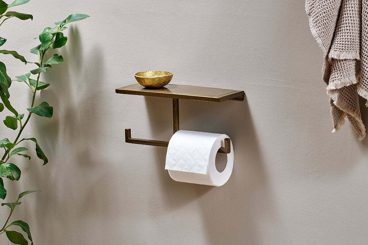The nkuku Mahi Wall Mounted Toilet Roll Holder in metal holds a gold dish above the toilet roll. A leafy plant is on the left, and textured beige towels hang on the right against a light wall.