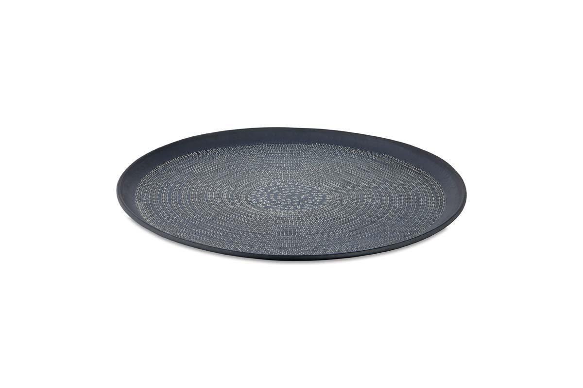 The Mahika Tray - Black by nkuku is a round, shallow aluminium tray with a black finish, raised edge, and hand-etched circular dotted pattern radiating from the center, shown on a plain white background.