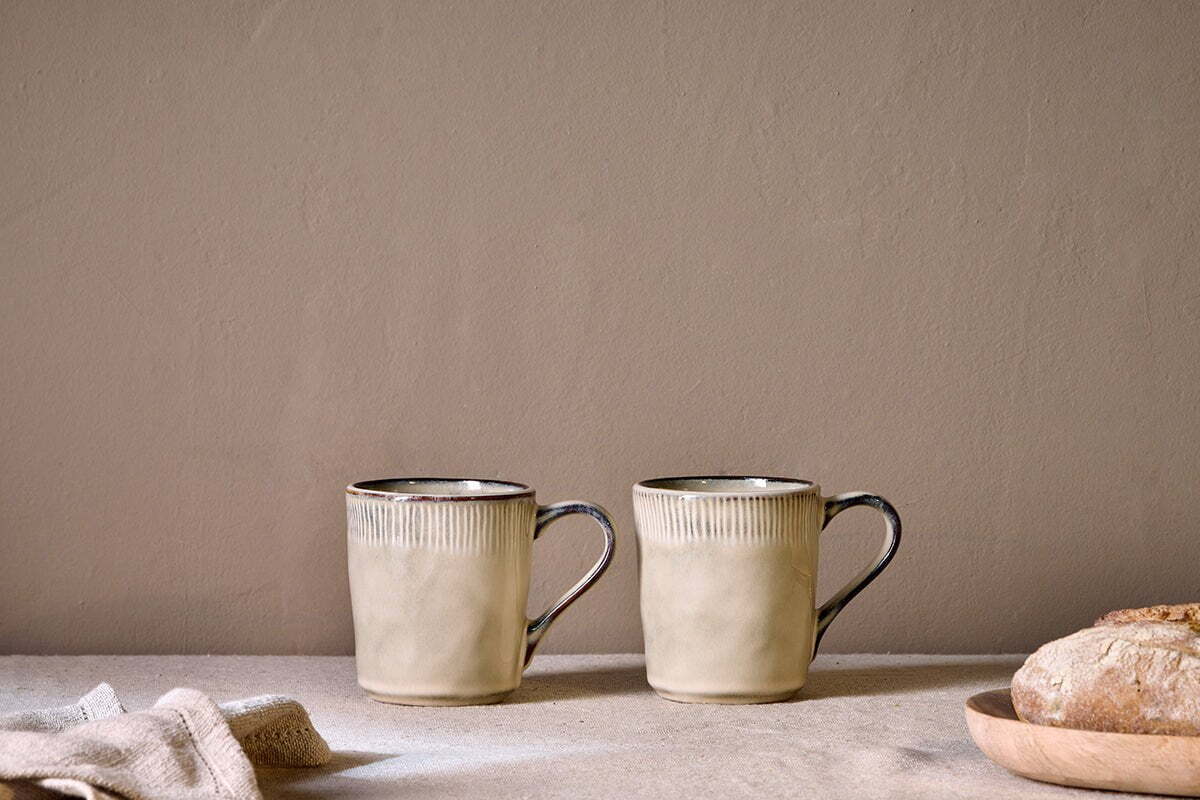 The nkuku Malia Mug - Cream (Set of 2) features textured rims and handles, styled side by side on a light surface with crusty bread and a folded cloth, set against a neutral wall for a minimalist, cozy vibe.