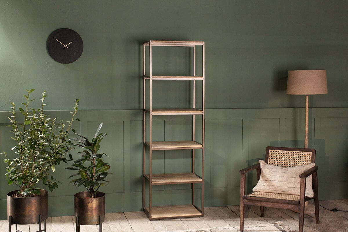 A minimalist room features the nkuku Marjori Mango Wood and Iron Standing Shelves - Narrow against a green wall, flanked by two potted plants, a black wall clock on the left, and a floor lamp with a wooden cane-back armchair on the right.