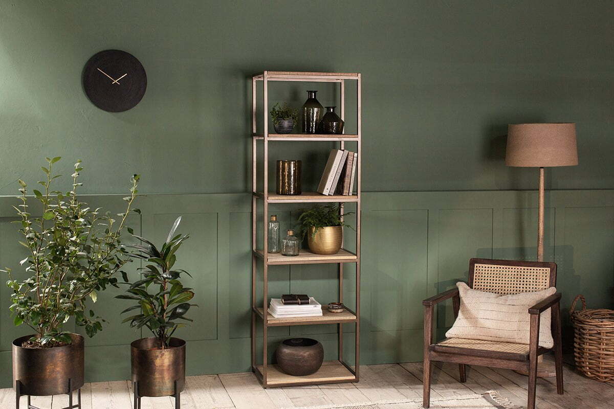 A cozy living room with a green wall, nkuku Marjori Mango Wood And Iron Standing Shelves - Narrow filled with books and decor, a black wall clock, two potted plants on stands, a wooden armchair with woven back, wicker basket, and floor lamp with beige shade.