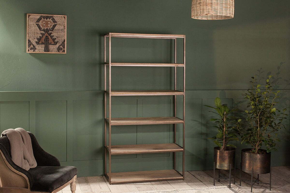 The Marjori Mango Wood And Iron Standing Shelves - Wide by nkuku stand empty against a green paneled wall, flanked by potted plants on black stands and a dark cushioned chair with a light throw. A woven pendant light and geometric artwork complete the look.