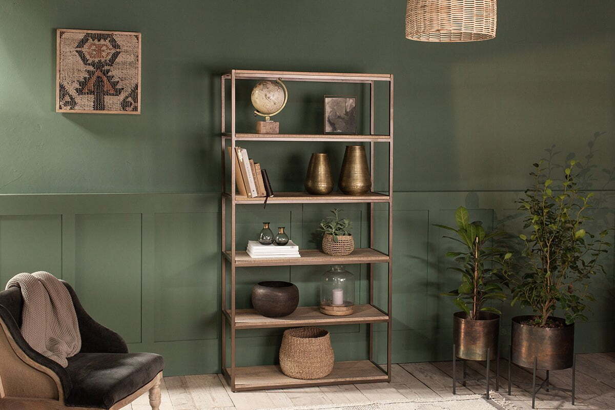 The nkuku Marjori Mango Wood And Iron Standing Shelves - Wide showcase books, vases, a globe, and plants against a green wall. Nearby potted plants, a plush gray chair, and a wicker pendant light create coziness beneath framed textile art.