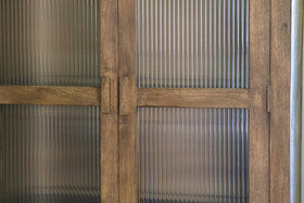 A close-up of nkuku’s Mendani Mango Wood & Fluted Glass Cabinet in a walnut stain shows vertical ribbed glass panels, visible grain on the light brown wood, simple rectangular handles, and stylish translucent fluted glass for added privacy.