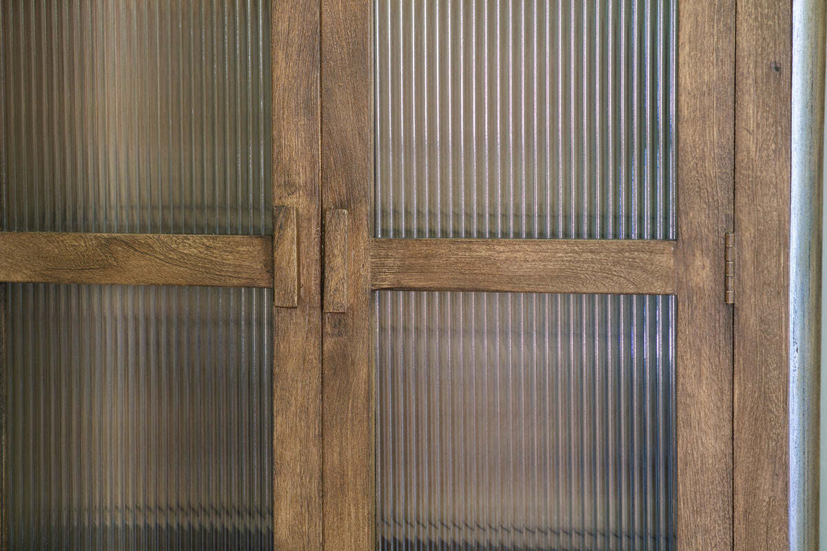 A close-up of nkuku’s Mendani Mango Wood & Fluted Glass Cabinet in a walnut stain shows vertical ribbed glass panels, visible grain on the light brown wood, simple rectangular handles, and stylish translucent fluted glass for added privacy.
