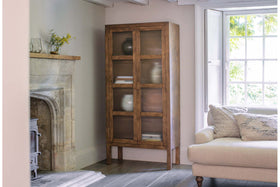 The nkuku Mendani Mango Wood & Fluted Glass Cabinet in walnut stain stands in a bright living room corner, displaying ceramic vases and books. Nearby, a beige sofa with textured pillows sits by the window, with a stone fireplace on the left wall.