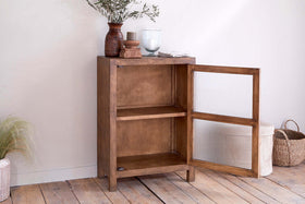 The nkuku Mendani Mango Wood & Fluted Glass Low Cabinet in a walnut stain stands on wooden floorboards, its glass-paneled door open to reveal two shelves. On top sit ceramic vases, wooden bowls, and a glass, with baskets and a plant nearby.