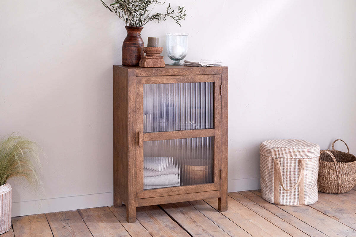 The Mendani Mango Wood & Fluted Glass Low Cabinet by nkuku in a walnut stain stands on a wooden floor, adorned with a vase, candle holder, and glass vase. Two woven baskets are positioned to its right.