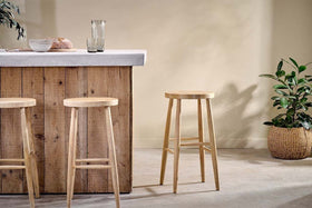 A rustic wooden bar with a concrete countertop features three nkuku Mengwi Mango Wood Round Bar Stools in natural. To the right sits a woven basket with a green plant, while a glass pitcher and two glasses are neatly arranged on the counter.