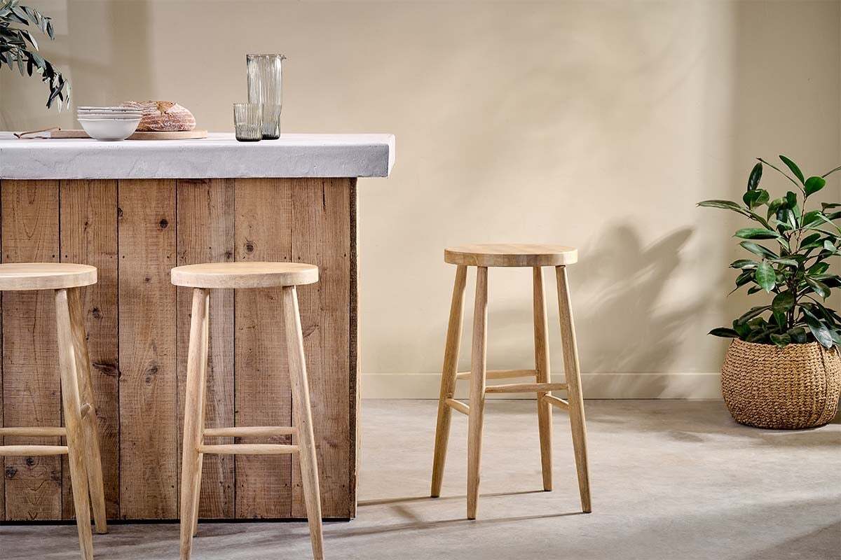 A rustic wooden bar with a concrete countertop features three nkuku Mengwi Mango Wood Round Bar Stools in natural. To the right sits a woven basket with a green plant, while a glass pitcher and two glasses are neatly arranged on the counter.