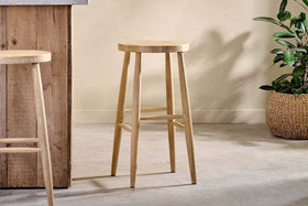 Two Mengwi Mango Wood Round Bar Stools by nkuku sit by a wood-paneled island, while a plant in a woven basket adds charm on the right.