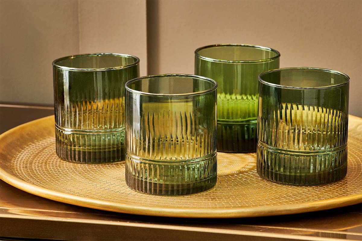 A set of four nkuku Mila Tumblers in Dark Emerald, with vertical ribbed and etched detail, are displayed on a round textured golden tray atop a reflective brown surface against a beige wall.