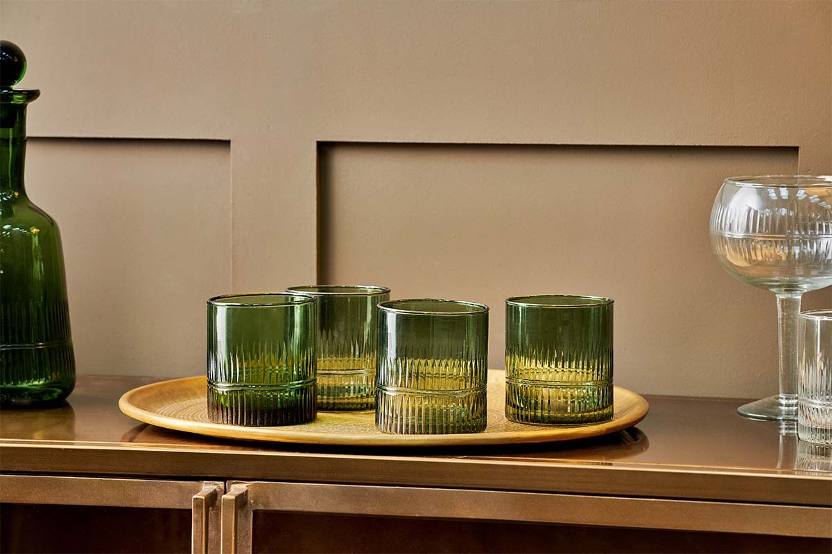 A round gold tray displays a set of four nkuku Mila Tumblers in Dark Emerald with ribbed glass, placed on a bronze sideboard. Also shown: a green decanter and two clear etched stemmed glasses against a beige paneled wall.