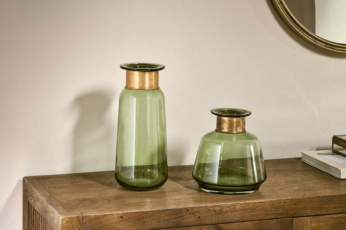 Two nkuku Miza Glass Vases in green, one tall and cylindrical, the other short and wide with copper rims, sit on a wooden surface. A round mirror and stacked books appear in the background against a light wall.