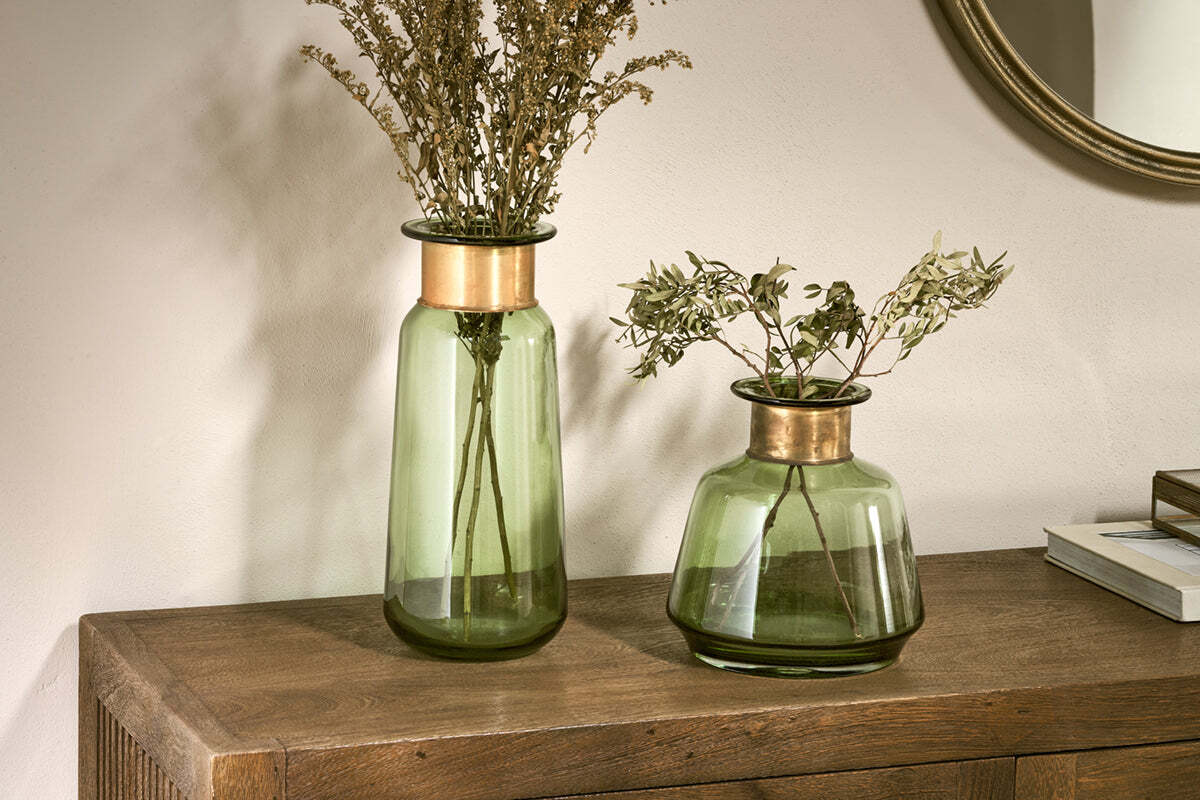 Two nkuku Miza Glass Vases in green sit on a wooden surface. The taller vase holds dried brown flowers, the shorter one has green leafy stems. A round mirror and stacked books are visible in the background.
