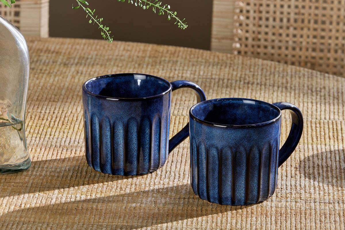 Two nkuku Moda Mugs - Deep Blue (Set of 2) with vertical ribbed patterns rest side by side on a woven beige tabletop, with a glass vase of greenery and a rattan chair back in the background.