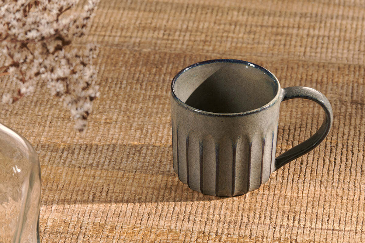 A set of two nkuku Moda Mug - Pewter with a grooved design and dark blue rim sits on textured wood, accompanied by blurred white dried flowers on the left.