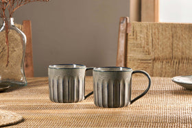 The nkuku Moda Mug - Pewter (Set of 2) features a grooved design and subtle pewter glaze, shown on a woven placemat with a chair and vase of dried branches softly lit by daylight from the window.