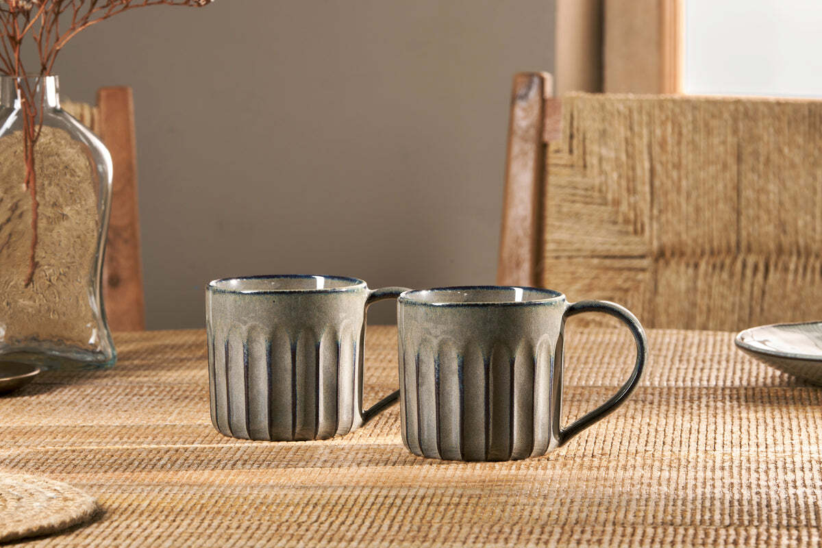 The nkuku Moda Mug - Pewter (Set of 2) features a grooved design and subtle pewter glaze, shown on a woven placemat with a chair and vase of dried branches softly lit by daylight from the window.