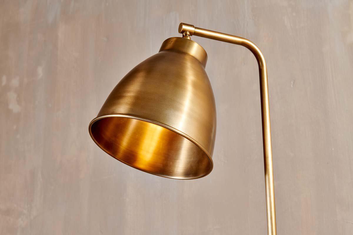 A close-up of the nkuku Muturi Floor Lamp - Antique Brass highlights its gold conical lampshade as a chic downlight, set on a sleek solid brass stand against a textured beige wall.
