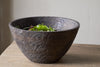 The nkuku Nyati Wooden Serving Bowl in dark brown (large) with a textured finish rests on a wooden table, partially filled with fresh green and purple leafy salad.
