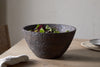 The nkuku Nyati Wooden Serving Bowl - Dark Brown - Large, filled with leafy greens, sits on a wooden table near a chair, beside a plate on a woven placemat and a light cloth.