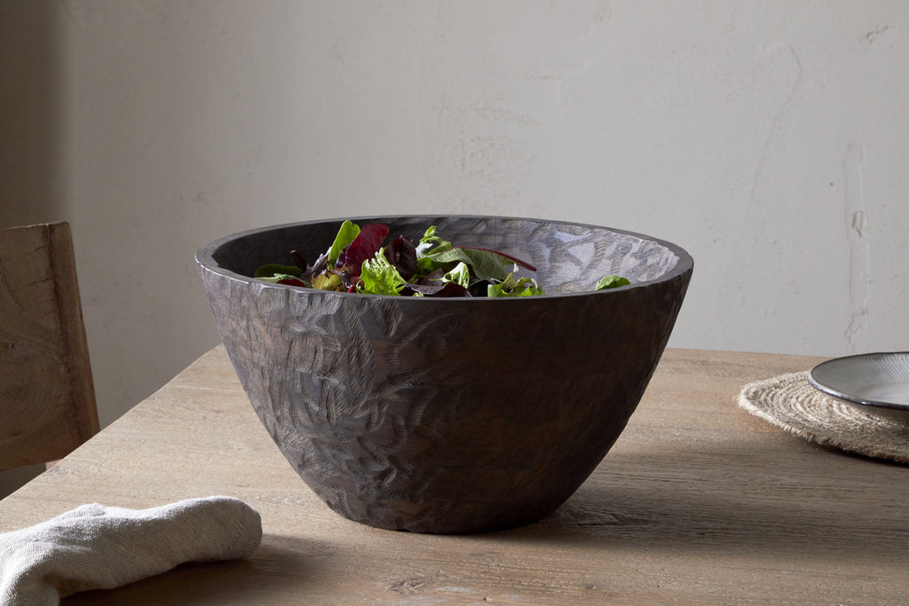 The nkuku Nyati Wooden Serving Bowl - Dark Brown - Large, filled with leafy greens, sits on a wooden table near a chair, beside a plate on a woven placemat and a light cloth.