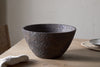 The large nkuku Nyati Wooden Serving Bowl in dark brown, handcrafted with a textured finish, sits on a light wooden table beside a beige cloth and woven placemat with a small plate, creating a minimal, rustic setting.