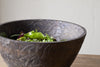 The nkuku Nyati Wooden Serving Bowl - Dark Brown - Large, handcrafted from mango wood, is filled with fresh mixed salad greens and placed on a wooden table against a light-colored wall.