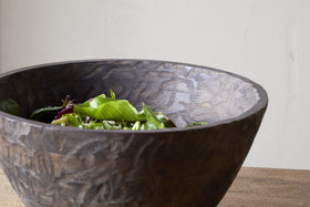 The nkuku Nyati Wooden Serving Bowl - Dark Brown - Large, handcrafted from mango wood, is filled with fresh mixed salad greens and placed on a wooden table against a light-colored wall.