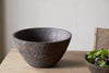 The nkuku Nyati Wooden Serving Bowl - Dark Brown - Large sits on a light wooden table beside a smaller bowl of green salad, with the back of a wooden chair visible against a plain, light wall.